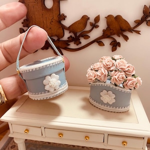 May include: Two miniature blue and white boxes with white lace trim and a white flower accent. One box is empty and the other box contains a bouquet of pink roses. The boxes are sitting on a white table with three drawers.