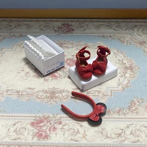 May include: Miniature red Mary Jane shoes with bow accents, displayed on a white box. A small white box with paper items and a red tool with black wheels are also present. The items are on a floral patterned surface.