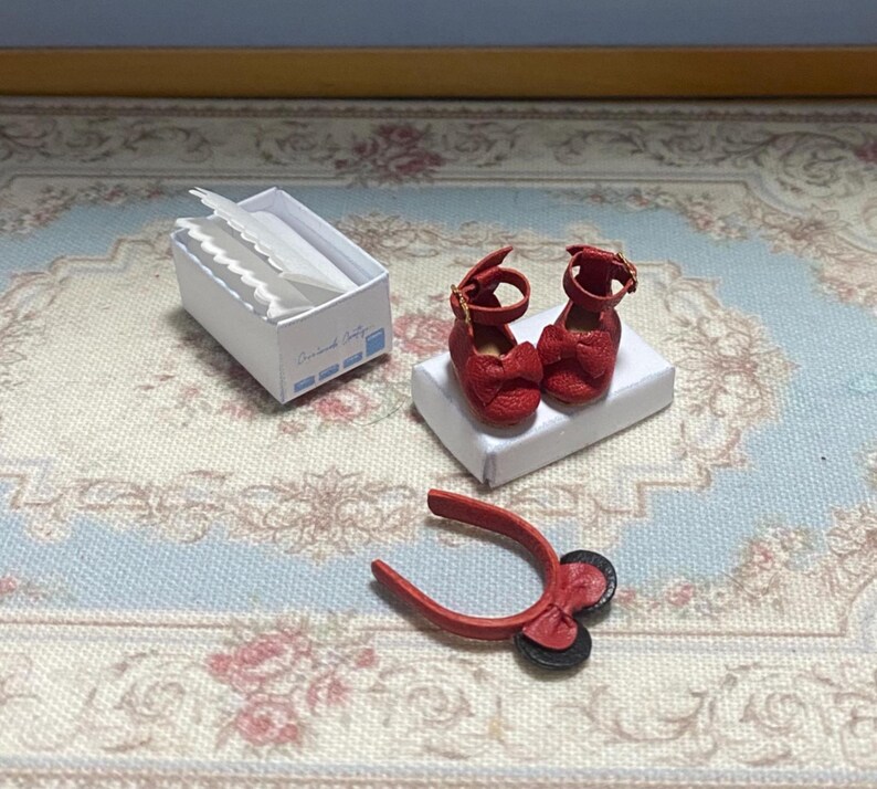 May include: Miniature red Mary Jane shoes with bow accents, displayed on a white box. A small white box with paper items and a red tool with black wheels are also present. The items are on a floral patterned surface.