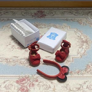 May include: Miniature red leather shoes with bow accents and a matching headband. A white box with paper inserts and a small white box with a blue logo are also present. The shoes have a strap with a gold buckle.