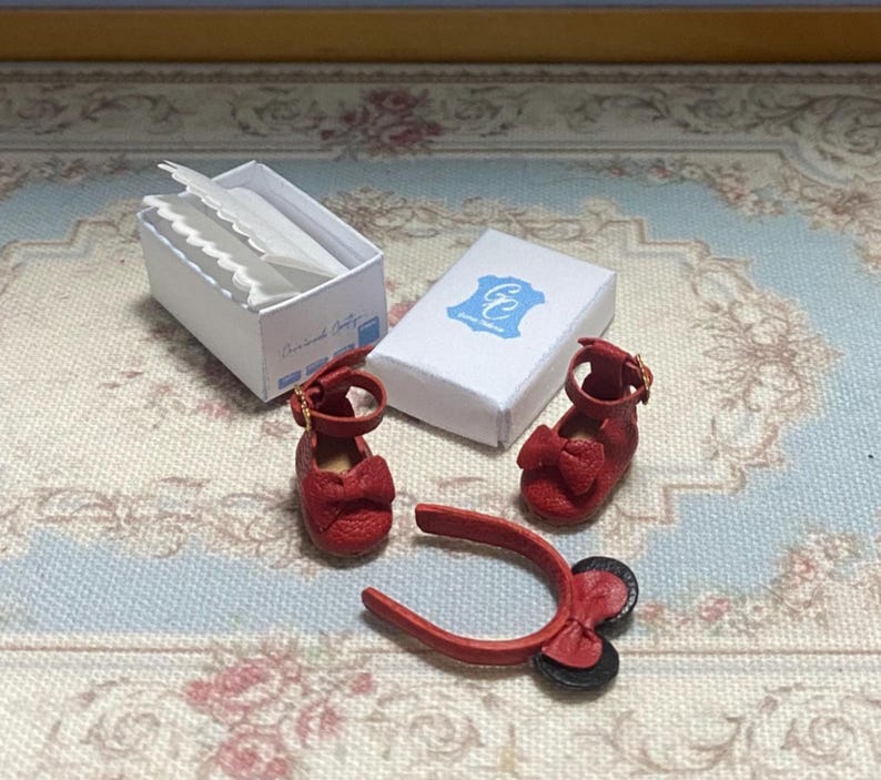 May include: Miniature red leather shoes with bow accents and a matching headband. A white box with paper inserts and a small white box with a blue logo are also present. The shoes have a strap with a gold buckle.