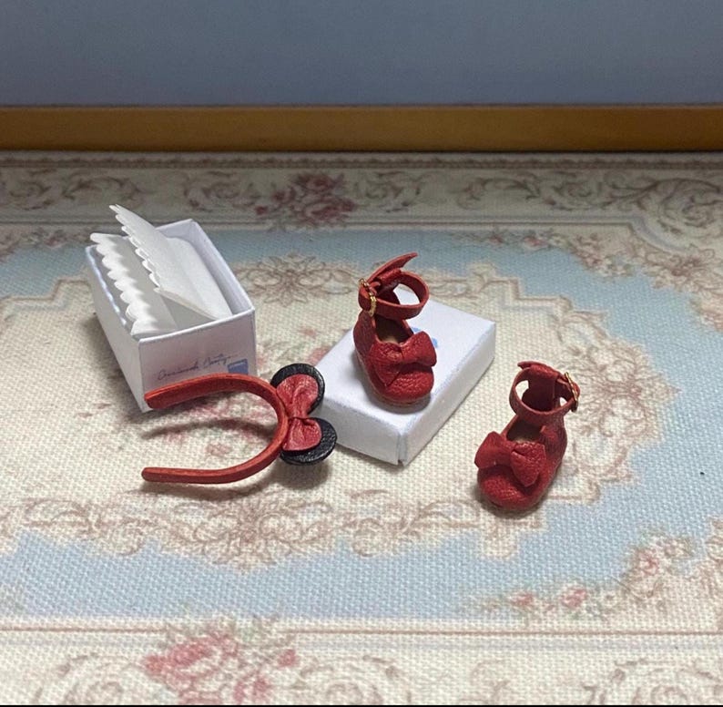 May include: Miniature red leather Mary Jane shoes with bow accents, a matching headband, and a small white box with tissue paper. The items are arranged on a floral patterned surface.
