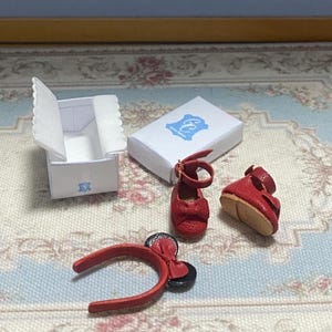 May include: Miniature red doll shoes, a headband with a bow, and two white boxes with blue logos. The shoes have ankle straps and bows. The headband has a bow and black circular accents. The boxes are open and closed.