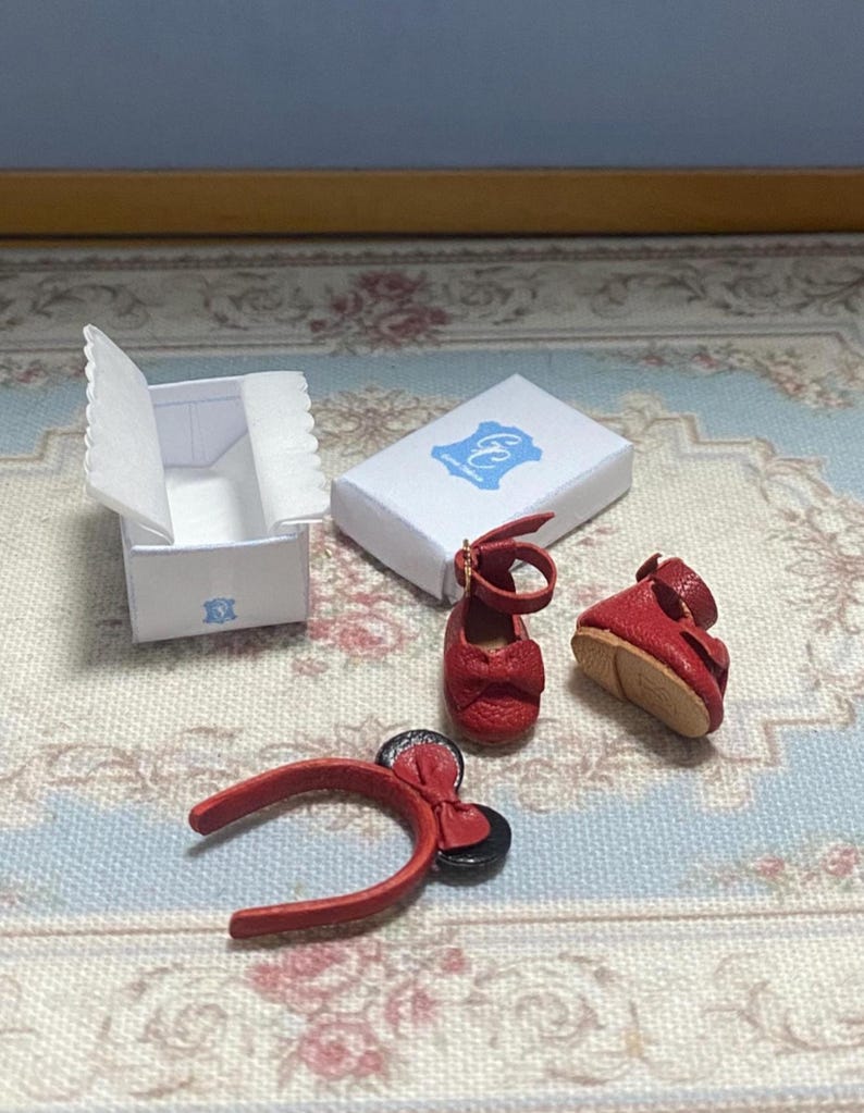 May include: Miniature red doll shoes, a headband with a bow, and two white boxes with blue logos. The shoes have ankle straps and bows. The headband has a bow and black circular accents. The boxes are open and closed.