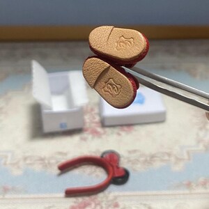May include: Miniature red shoes with tan soles, held by tweezers. The soles feature a decorative embossed design. A red headband with a bow and a small white box are also visible in the image.