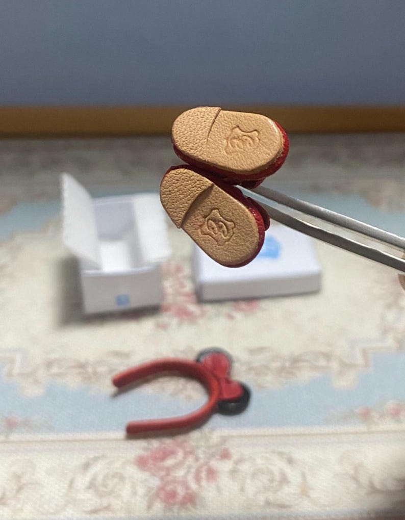 May include: Miniature red shoes with tan soles, held by tweezers. The soles feature a decorative embossed design. A red headband with a bow and a small white box are also visible in the image.