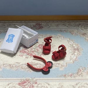 May include: Miniature red leather shoes with bows and straps, alongside a matching headband and a white box with a blue logo. The shoes are designed with intricate details, suggesting a doll or collectible item.