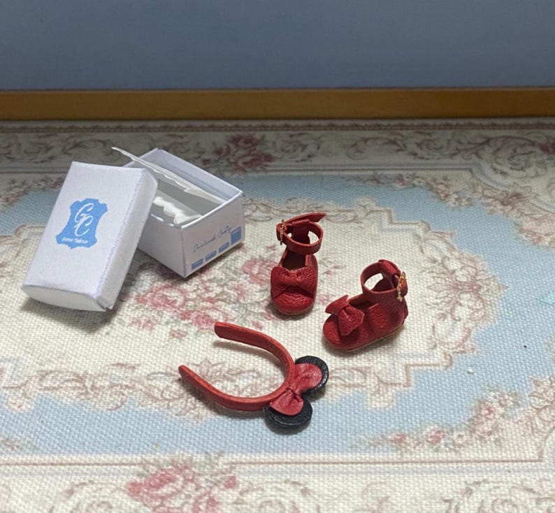 May include: Miniature red leather shoes with bows and straps, alongside a matching headband and a white box with a blue logo. The shoes are designed with intricate details, suggesting a doll or collectible item.
