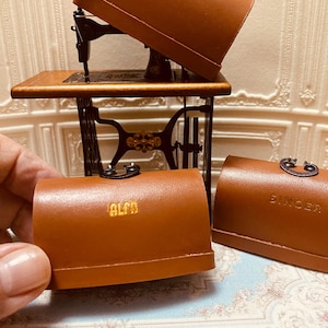 Può includere: Tre custodie per macchine da cucire in miniatura con i nomi dei marchi "SIGMA", "ALFA" e "SINGER" stampati su di esse. Le custodie sono in pelle marrone e hanno un aspetto vintage.