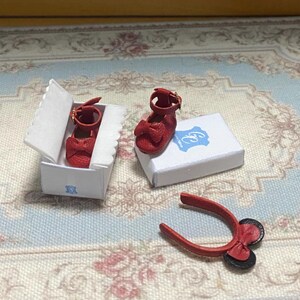 May include: Miniature red shoes with bow accents, a matching headband, and a white box with a blue logo. The shoes have a strap with a gold buckle. The headband has a bow and two black circles.
