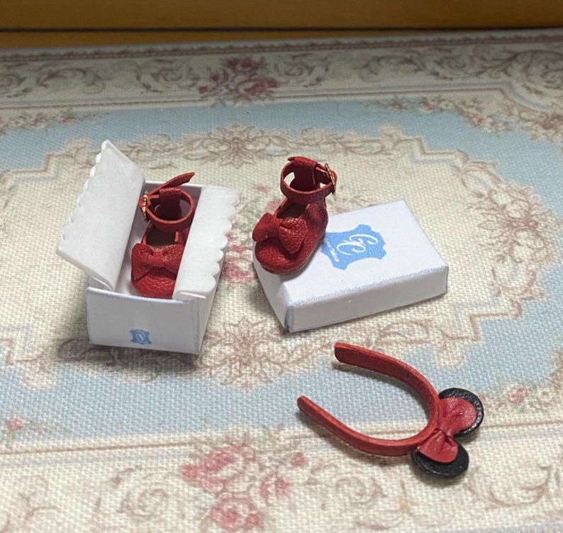 May include: Miniature red shoes with bow accents, a matching headband, and a white box with a blue logo. The shoes have a strap with a gold buckle. The headband has a bow and two black circles.