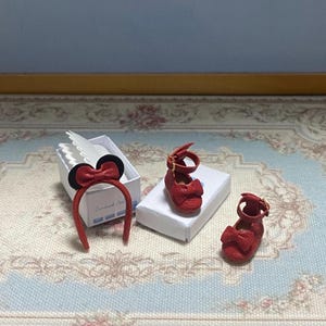 May include: A set of miniature red doll shoes with bow accents and a matching headband with a bow and black ears. The items are displayed on a patterned surface, suggesting a children's or doll-themed product.