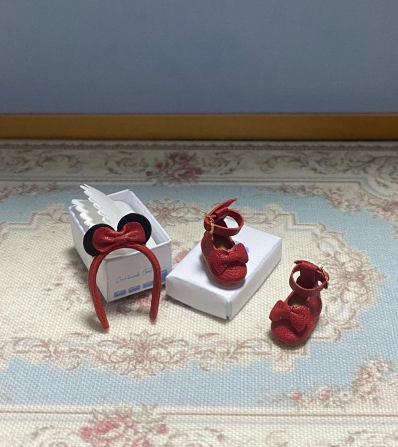 May include: A set of miniature red doll shoes with bow accents and a matching headband with a bow and black ears. The items are displayed on a patterned surface, suggesting a children's or doll-themed product.