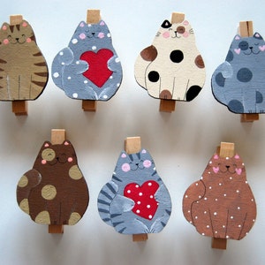 Può includere: Set di otto mollette di legno dipinte con diversi disegni di gatti. I gatti indossano tutti colori e motivi diversi, tra cui marrone, grigio, bianco e nero. Alcuni dei gatti tengono cuori.