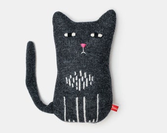 Puede incluir: Un peluche de gato gris oscuro hecho a mano con detalles bordados en blanco. El gato tiene una nariz rosa, ojos negros y blancos y una cola larga. Una etiqueta roja con texto blanco está adjunta al costado.