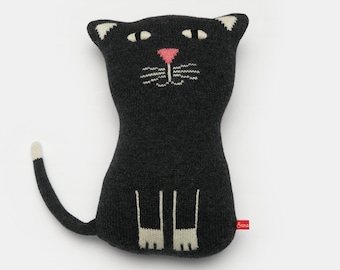 Puede incluir: Una almohada de gato de punto negro con detalles blancos en las patas y la cola. El gato tiene la nariz rosa y los ojos blancos. La almohada está hecha por Boon.