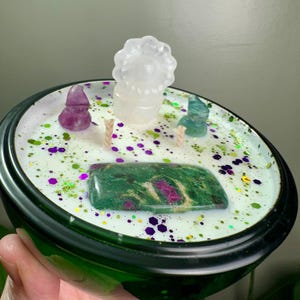 May include: A green glass candle holder filled with white wax, decorated with crystals and glitter. The candle features a large green and red stone, a clear crystal, and two small crystal figurines. The candle is held in a hand.