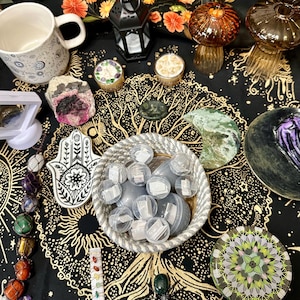 May include: A woven basket filled with clear plastic capsules containing small white paper scrolls, surrounded by a black cloth featuring a gold tree design, a white mug, a black lantern, crystal stones, a hamsa hand dish, and a spider figurine.