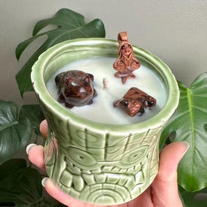 May include: A green tiki-style candle holder filled with white wax. Three brown frog and gnome figurines are placed in the wax. The candle holder is held by a person's hand.
