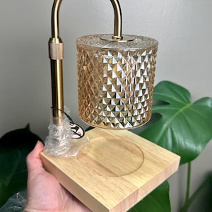 May include: A candle warmer lamp featuring a gold-toned metal arm and a cylindrical, amber-colored glass shade with a diamond-cut textured pattern. The lamp is mounted on a square, light-colored wooden base with a circular indentation in the center.