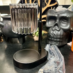 May include: A black candle warmer lamp featuring a cylindrical, fluted glass shade suspended from a curved metal arm. The base is oval-shaped with a flat surface for placing a candle. A power cord with a control switch is wrapped in plastic.