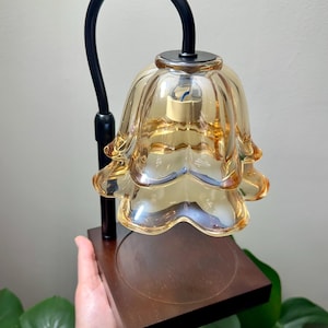 May include: A candle warmer lamp featuring a tiered, amber-colored glass shade shaped like a flower. The lamp has a matte black curved metal arm attached to a square, dark brown wooden base with a circular indentation for a candle.