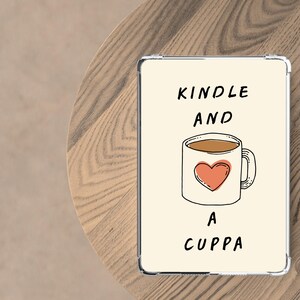 May include: A clear e-reader case with the text "KINDLE AND A CUPPA". The case has a drawing of a coffee mug with a heart. The case is on a wooden table, perfect for reading with a hot drink.