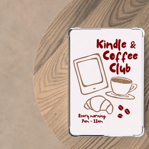 May include: A white sign featuring the text "Kindle & Coffee Club" in maroon script, accompanied by illustrations of a tablet, coffee cup, croissant, and coffee beans. The sign also includes the text "Every morning 7am - 11am."