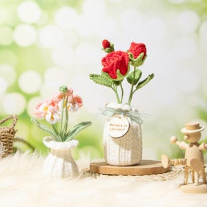 Op de afbeelding: Gehaakte bloemen in witte vazen. Eén vaas bevat rode rozen met groene bladeren, de andere roze en witte bloemen. Een houten label op de rozenvaas luidt "Believe in yourself".