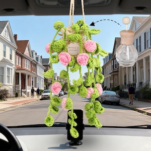 Może przedstawiać: Szydełkowy dodatek do samochodu z wiszącym wzorem rośliny, różowymi kwiatami i zielonymi liśćmi. Zawiera mały drewniany dyfuzor i przezroczystą szklaną butelkę. Widoczny jest napis "Believe in yourself".