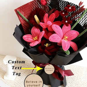 Puede incluir: Un ramo de flores rojas y rosas envuelto en papel negro, atado con una cinta burdeos. Una etiqueta de madera con el texto "Believe in yourself" está adjunta. El ramo incluye varios tipos de flores.