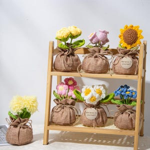 Puede incluir: Una estantería de madera exhibe varias flores de ganchillo en sacos de yute. Las flores incluyen variedades amarillas, rosas, blancas y azules, con hojas verdes. Algunos sacos tienen una etiqueta que dice "Believe in yourself".