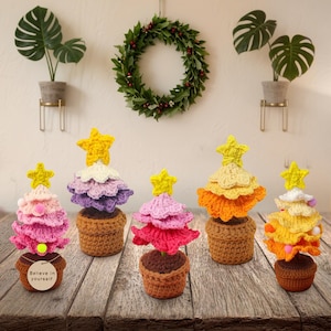 May include: A collection of five handmade crochet Christmas trees in various colors, including pink, purple, and yellow, each topped with a yellow star. The trees are potted in brown crochet pots and arranged on a wooden surface.