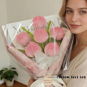 May include: A bouquet of pink tulips wrapped in clear cellophane. The stems are green, and the flowers are tied with a white ribbon and a tag that says "Believe in yourself". The bouquet is held by a person.