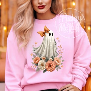 May include: A pink sweatshirt with a white ghost wearing an orange bow and surrounded by orange and white flowers. The text "Pudge McFluff" is in a white circle with a dashed border.
