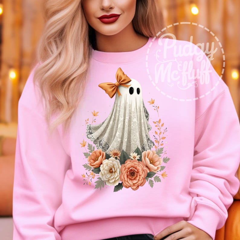 May include: A pink sweatshirt with a white ghost wearing an orange bow and surrounded by orange and white flowers. The text "Pudge McFluff" is in a white circle with a dashed border.