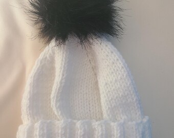 May include: A white knit beanie with a folded brim and a large, fluffy black pom-pom on top. The hat is made of a soft, textured yarn, perfect for cold weather.