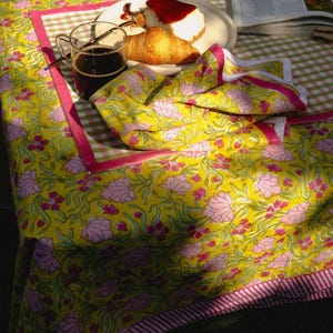May include: A yellow floral tablecloth with pink accents and a pink striped border. A plate with a slice of cheesecake and a croissant sits on the table, along with a cup of coffee and a folded napkin.
