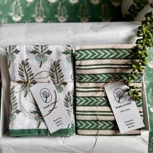 May include: Two sets of block print textiles, one with a floral design and the other with a chevron pattern, tied with twine and tags. The textiles are white and green, and are labeled as napkins and placemats.
