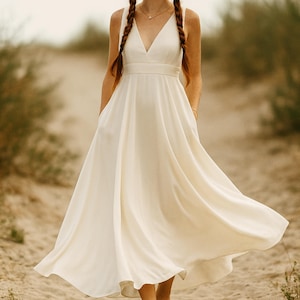 Peut inclure: Une robe sans manches de couleur ivoire avec un décolleté en V et une jupe fluide. La robe a un corsage ajusté et une taille définie. Le modèle se tient sur un chemin sablonneux, la robe bougeant doucement dans le vent. La robe a des poches.