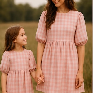 May include: Two matching pink gingham dresses. One is worn by a young girl, the other by an adult. Both dresses have short, puffed sleeves and a gathered waist. The girl and adult are holding hands.