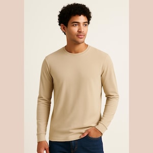 Pode incluir: Uma camisa de manga comprida de cor bege lisa, com gola redonda. A camisa tem um design simples e clássico com uma textura canelada. O modelo usa jeans azuis. A camisa é feita de um material macio e confortável.