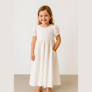 Könnte beinhalten: Ein helles Kleid mit einem Muster aus kleinen rosa Herzen. Das Kleid hat kurze, gepuffte Ärmel, eine geraffte Taille und Taschen. Das Mädchen lächelt und trägt das Kleid. Das Kleid hat eine Midi-Länge.