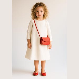 May include: A child in a cream-coloured dress, red shoes, and a small red handbag with a long strap. The background is plain white. The dress has long sleeves. The shoes and bag are a vibrant red.