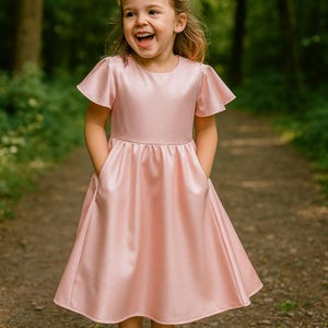 以下が含まれることがあります： 短いフレアスリーブとフレアスカートが付いた、明るいピンク色のサテンのドレス。 ドレスは丸いネックラインとポケットが付いています。 女の子は笑顔で、森の中の小道に立っています。