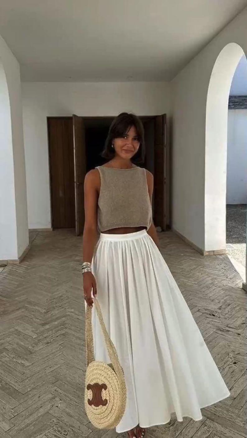 May include: A woman wearing a brown sleeveless top and a white pleated skirt. She is carrying a round straw bag with a brown leather logo.