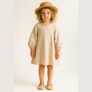 Op de afbeelding: Een jong kind draagt een beige linnen jurk met lange, pofmouwen en een V-hals. Het kind draagt ook een strohoed en beige schoenen. De jurk heeft een eenvoudig, elegant ontwerp.