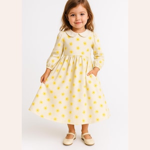 May include: A light yellow dress with a floral pattern of yellow flowers. The dress has long sleeves, a Peter Pan collar, and pockets. The girl is wearing cream-colored Mary Jane shoes.