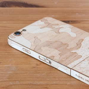 May include: A wooden phone case with a camouflage pattern. The case is light brown with a white camouflage design. The case has a cutout for the camera lens and a small opening for the charging port.