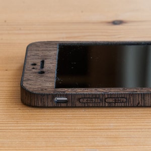 May include: A dark brown wooden phone case with a black screen and a silver charging port. The case has a wood grain pattern and is designed to fit a smartphone.